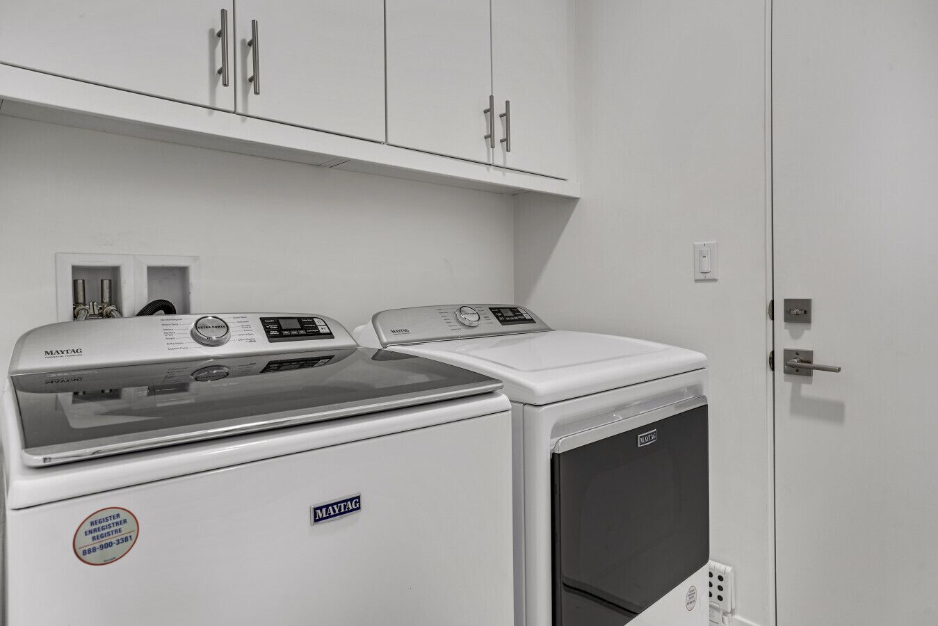 Une buanderie équipée de deux machines à laver et d’un sèche-linge Maytag, avec des placards blancs au-dessus et une porte dotée d’une poignée.