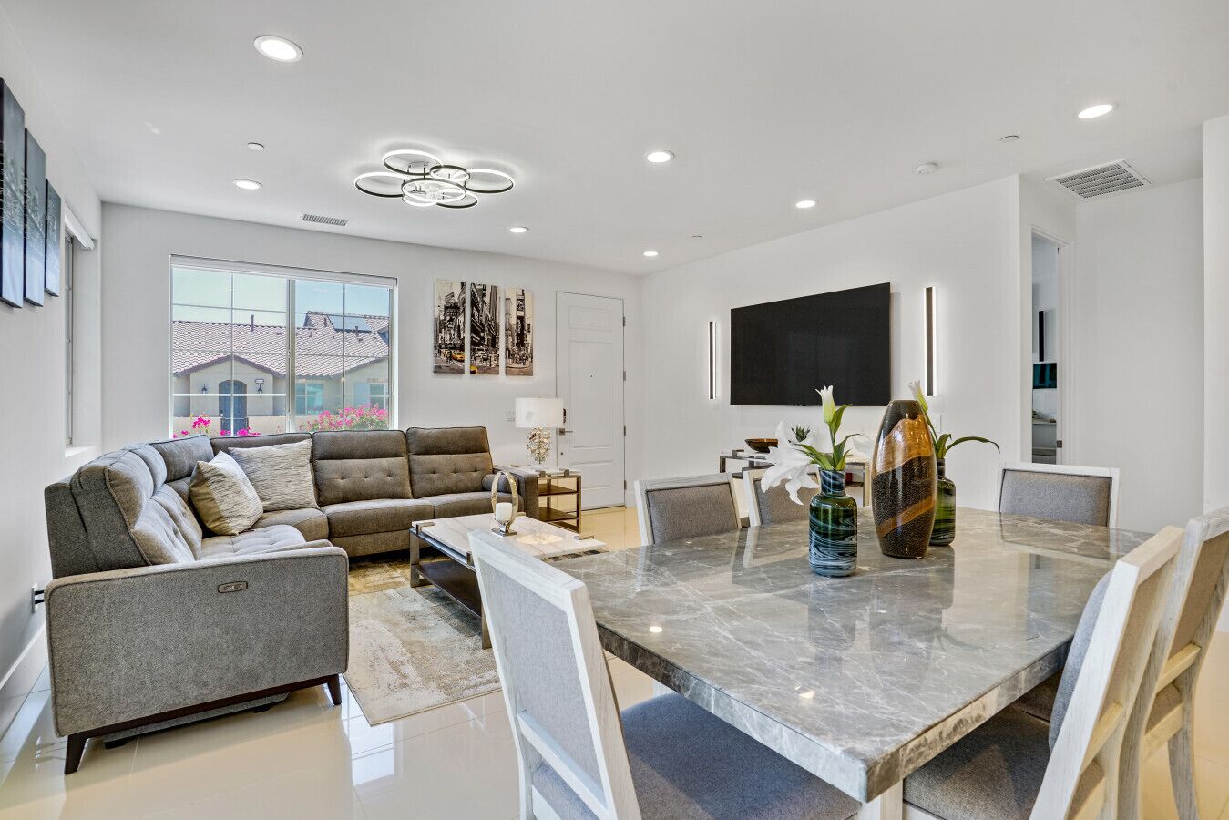 A modern living room with a grey sofa, a dining table with chairs, and a large window with a view of buildings.