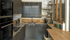 Eine moderne Küche mit Einbauofen, Spüle und einer Holzarbeitsplatte. Regale mit Gläsern und ein Fenster mit Jalousien.