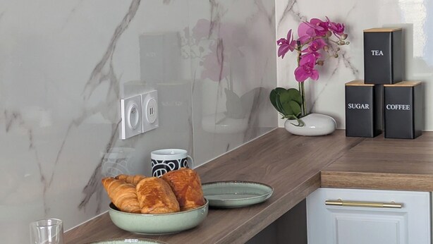 Une cuisine avec un dosseret en marbre, un plan de travail en bois, un vase rempli de fleurs et des boîtes noires étiquetées « SUCRE » et « CAFÉ ».