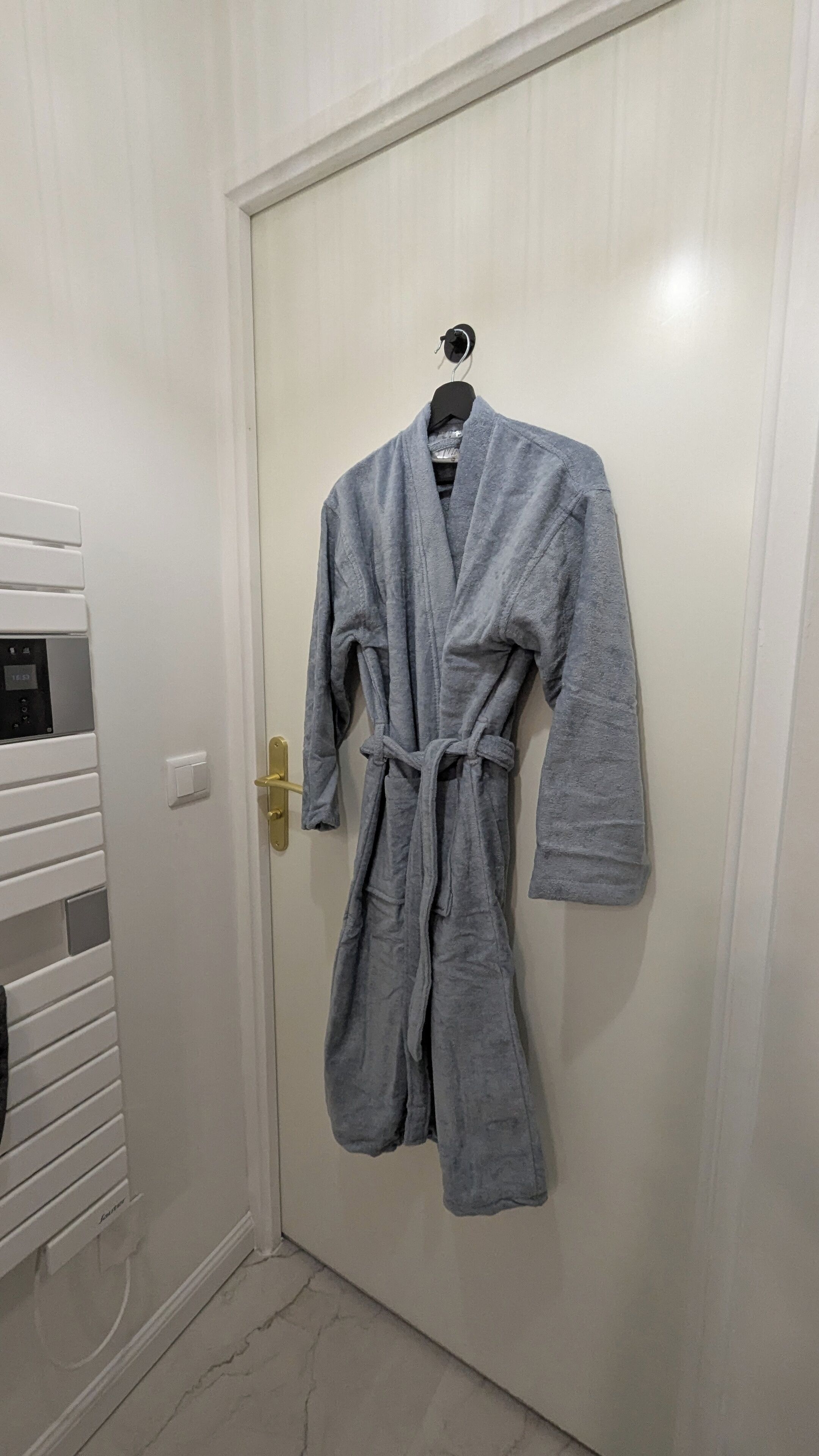 Une blouse grise est accrochée à une porte, à côté d’un thermostat numérique et d’un interrupteur d’éclairage fixés au mur.