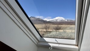 Een raam met uitzicht op een berg met sneeuw, een heldere lucht en een droog landschap met schaarse vegetatie.