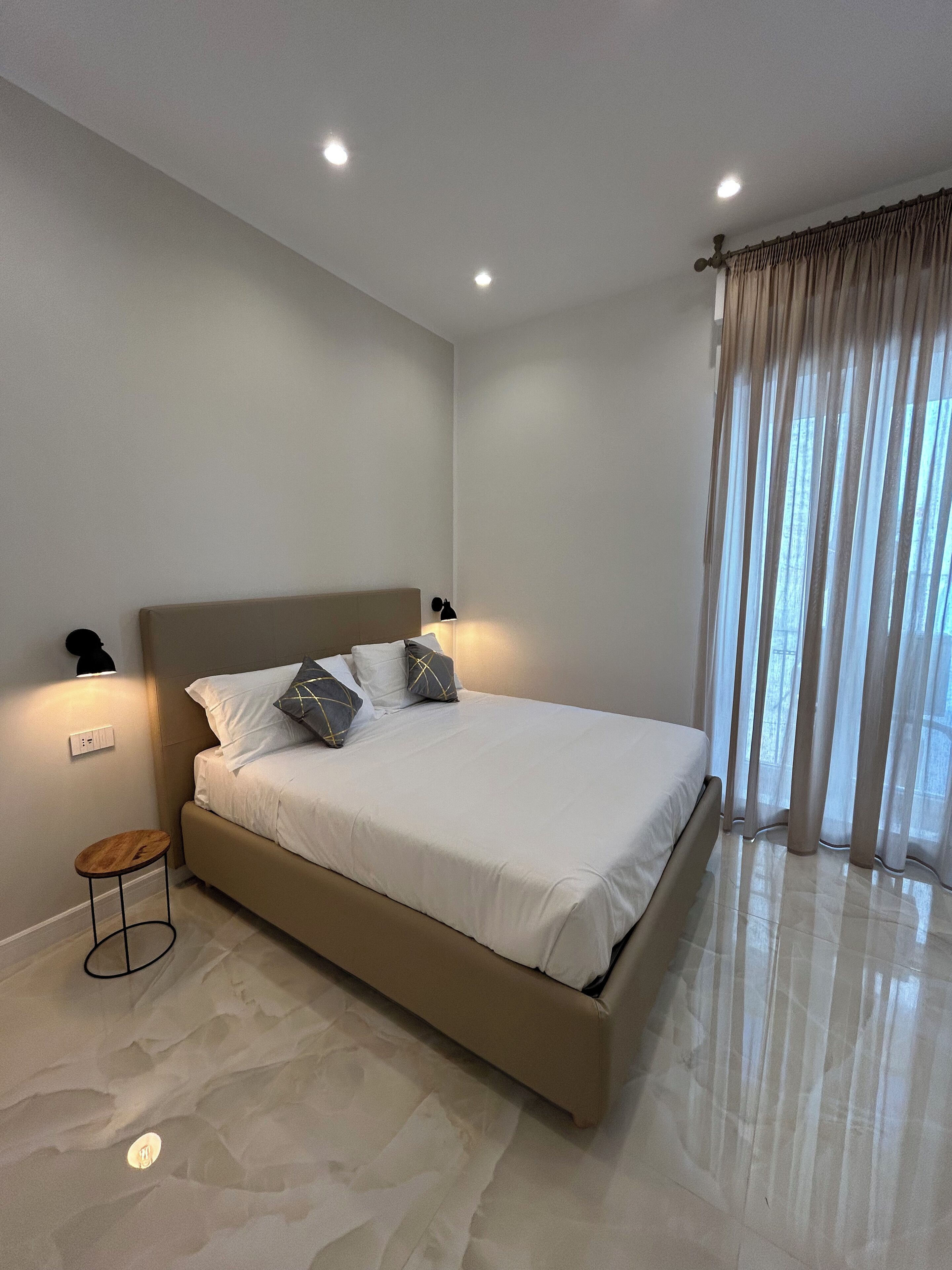 Un dormitorio moderno con una cama grande, dos lámparas de noche, un taburete pequeño y un ventilador de techo.