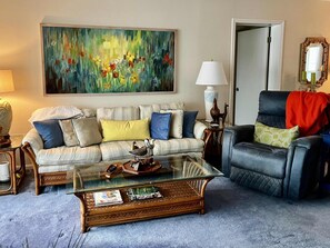 A living room with a sofa, armchair, coffee table, and a painting on the wall. - Island Reef #501 Beachfront Condo (Fort Myers Beach)