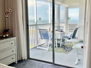 A room with a sliding glass door leading to a balcony with a view of the sea. There is a white wicker dresser, a potted plant, and a table with chairs.