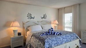 Une chambre à coucher avec un lit, deux tables de chevet avec des lampes, une fenêtre avec des rideaux et des œuvres d’art murales.