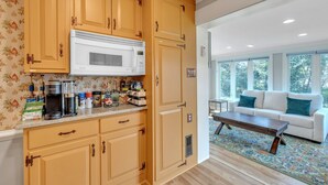 Een keuken met houten kastjes, een magnetron en een koffiezetapparaat. In de aangrenzende woonkamer staat een witte bank en ligt een vloerkleed met een patroon.