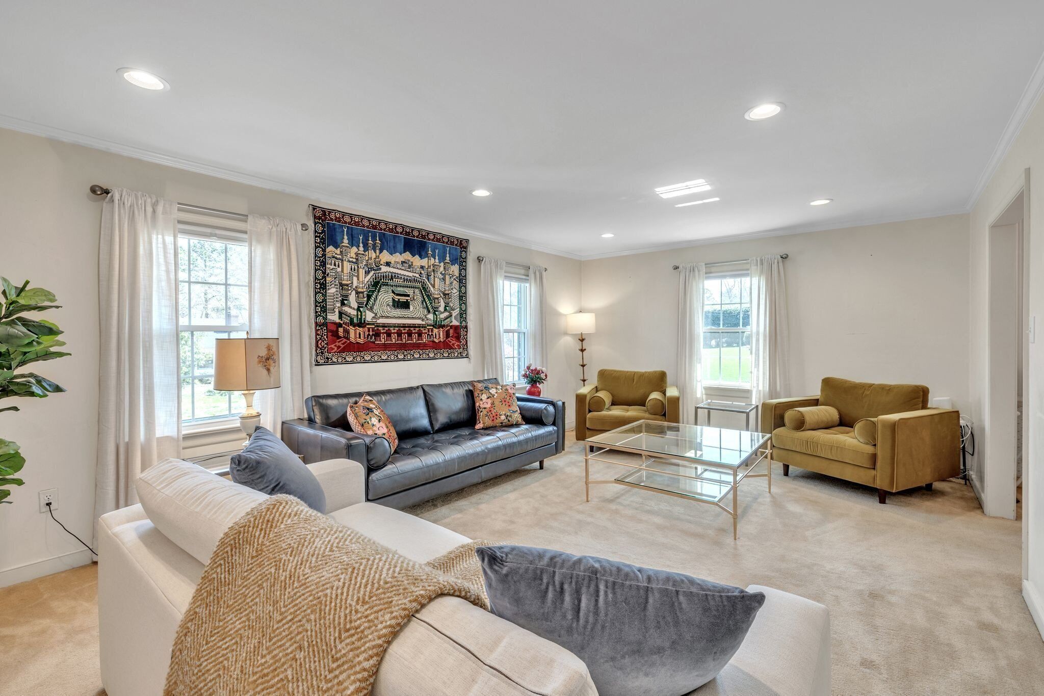 A spacious living room with a large sofa, a coffee table, and two armchairs. There is a large tapestry on the wall and a window with curtains.