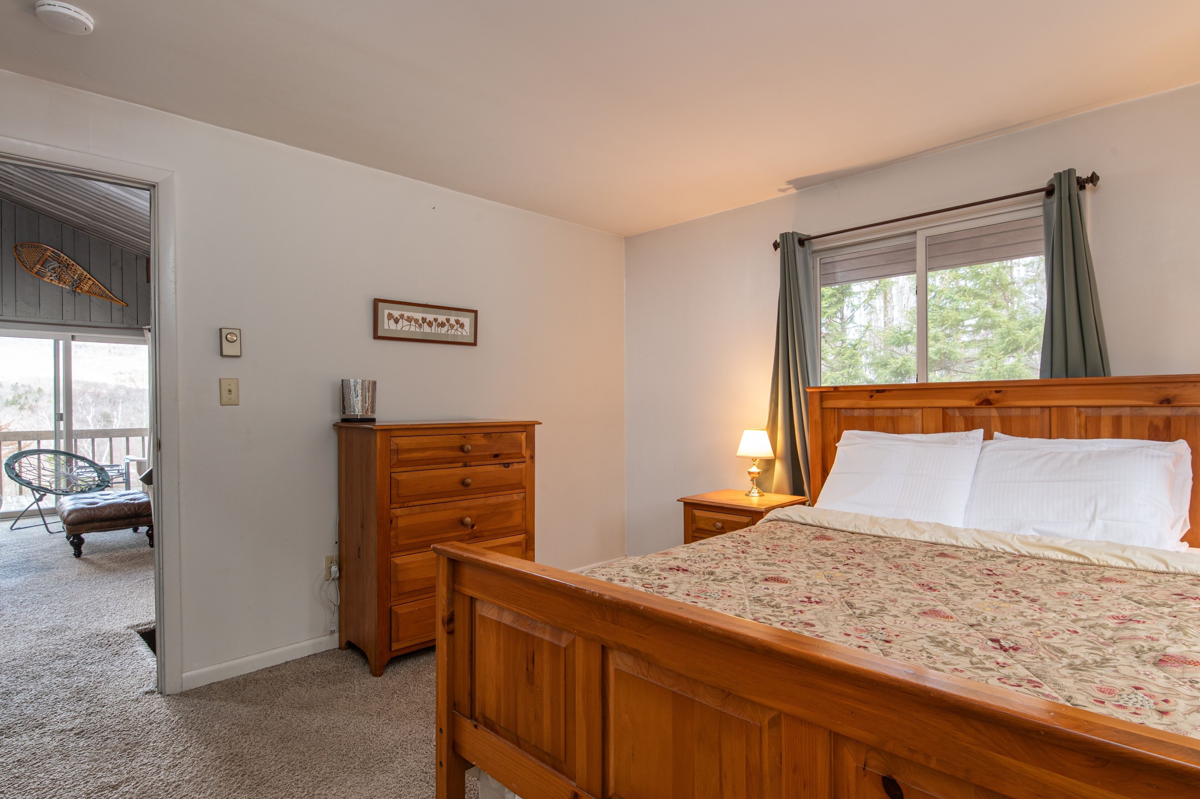 A bedroom with a wooden bed, a nightstand with a lamp, a dresser, and a window with curtains.
