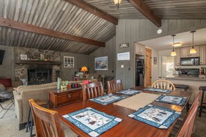 A dining area with a wooden table, chairs, and a fireplace. There is a kitchen visible in the background. - Bear Hill Cabin 4 Bedroom Cabin (Ludlow)