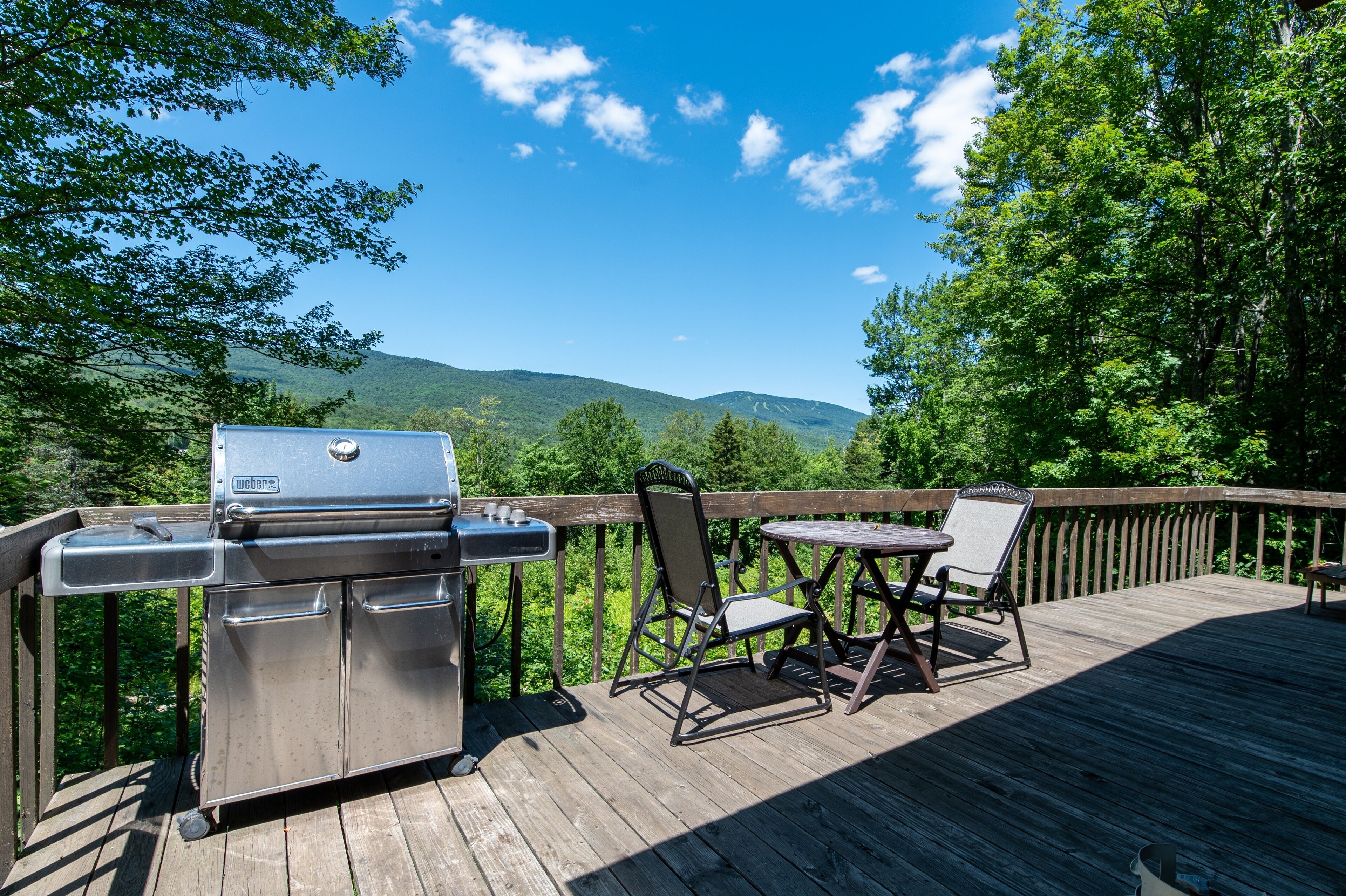 Une terrasse en bois équipée d’un barbecue, de deux chaises et d’une table, donnant sur une zone boisée avec des montagnes en arrière-plan.