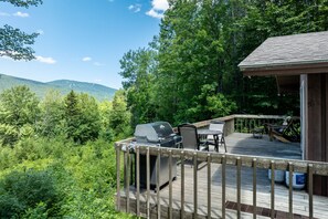 A wooden deck with a grill, chairs, and a table, surrounded by lush greenery and mountains. - Bear Hill Cabin 4 Bedroom Cabin (Ludlow)
