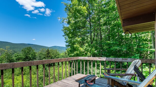 Eine Holzterrasse mit einer Bank und einem Tisch, mit Blick auf ein bewaldetes Gebiet und Hügel im Hintergrund.