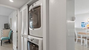 A laundry room with a front-loading washer and dryer, a chair, and a table with a painting. - Kihei Beach, #109 2 Bedroom Condo (Kihei)