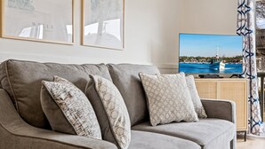 A grey sofa with patterned cushions, a flat-screen TV displaying a coastal scene, and a patterned curtain.