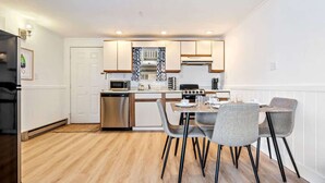 A compact kitchen with modern appliances, a dining area with grey chairs, and a white door in the background.