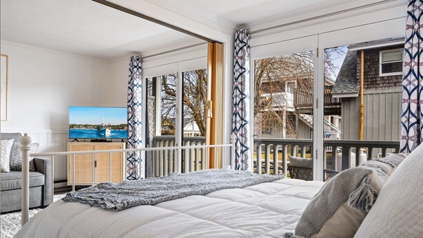 A bedroom with a large bed, a television, a sofa, and a view of a wooden deck and a boat through the window.