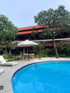 A swimming pool with lounge chairs and umbrellas, surrounded by lush greenery and a building with a red-tiled roof. - Uns Hotel - Bali (Kuta)