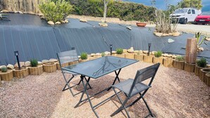 Een terras buiten met een tafel en stoelen, omringd door decoratieve plantenbakken en een veld met zonnepanelen.