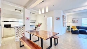 A modern kitchen with white cabinets, a wooden dining table with benches, pendant lighting, and a living area with a blue sofa and artwork.