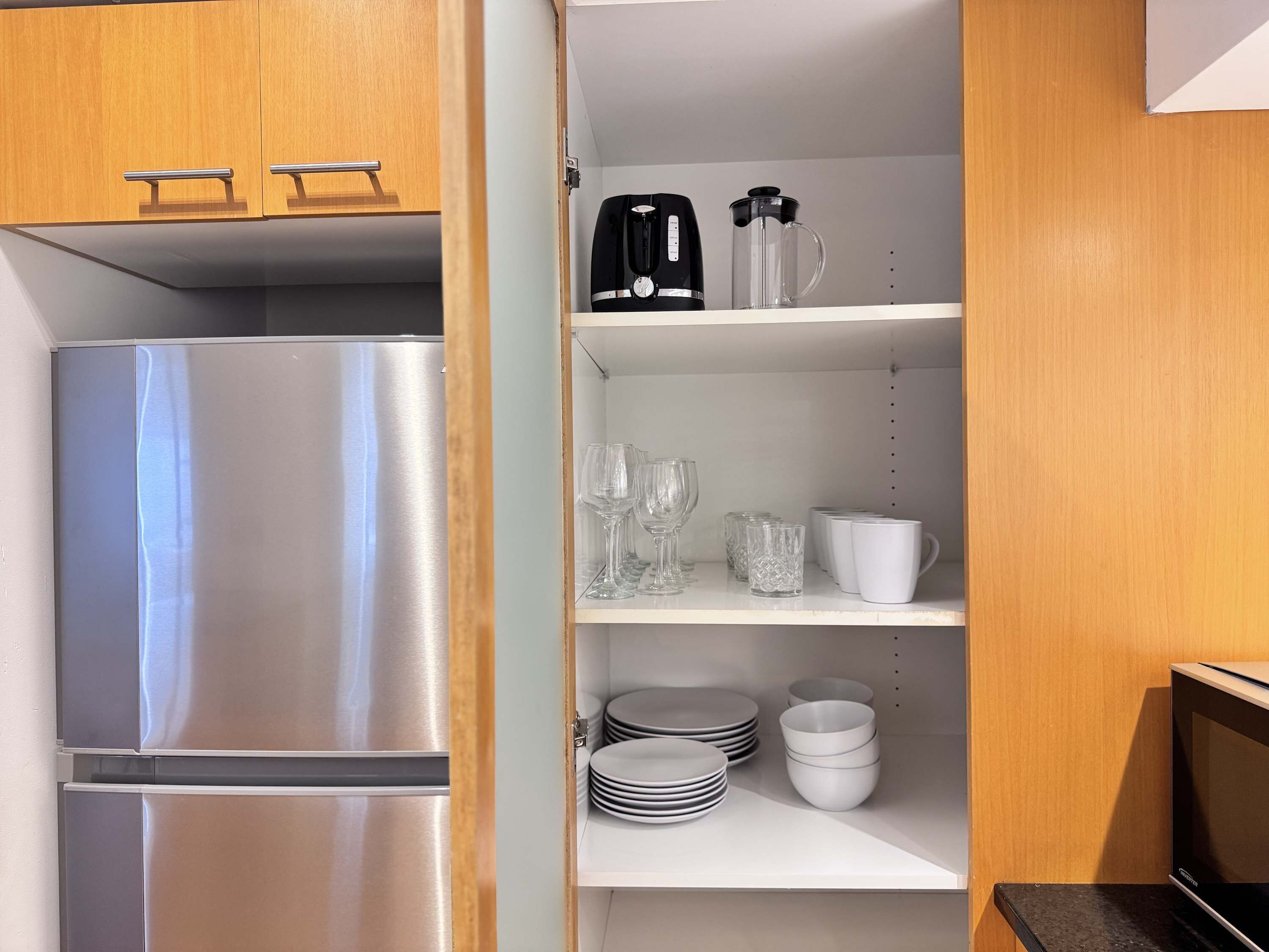 Une cuisine compacte équipée d’un réfrigérateur en acier inoxydable, d’une machine à café, ainsi que de divers verres et plats rangés sur des étagères.