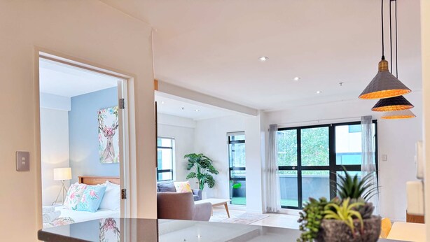 A modern hotel room with a large bed, a sitting area with a sofa, a dining table, and a view of the outdoors through large windows. - Heart of Auckland 2BR Near Queen St & Skytower (Auckland)