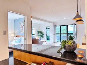 A modern hotel room with a large bed, a sitting area with a sofa, a dining table, and a view of the outdoors through large windows. - Heart of Auckland 2BR Near Queen St & Skytower (Auckland)