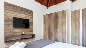 A modern bedroom with a wooden wall-mounted TV, a bed with a grey blanket, and a pair of rolled towels.