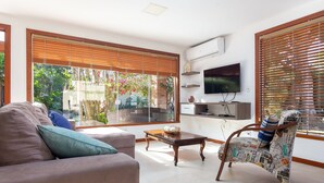 A living room with a sofa, coffee table, and a patterned armchair. There is a television on the wall and large windows with blinds.