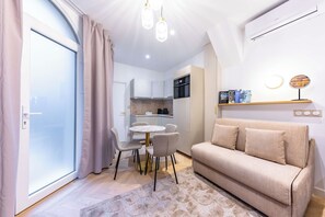 A modern living room with a sofa, dining table, and chairs. There is a kitchen area with cabinets and a stove. The room has a large window with curtains.