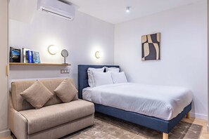 A bedroom with a bed, a sofa, a bedside table with books, a wall-mounted light, and a wall art piece.