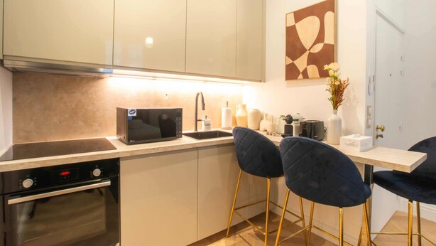 A modern kitchen with a built-in oven, a microwave, and a sink. There are two dark blue chairs with gold legs and a small table with a vase and a box.