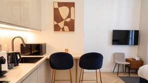 A modern kitchen with a microwave, kettle, and coffee maker. There are two bar stools and a chair. A flat-screen TV is mounted on the wall.