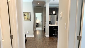 Un couloir au plancher de bois menant à une cuisine équipée d’un réfrigérateur et d’un luminaire suspendu.
