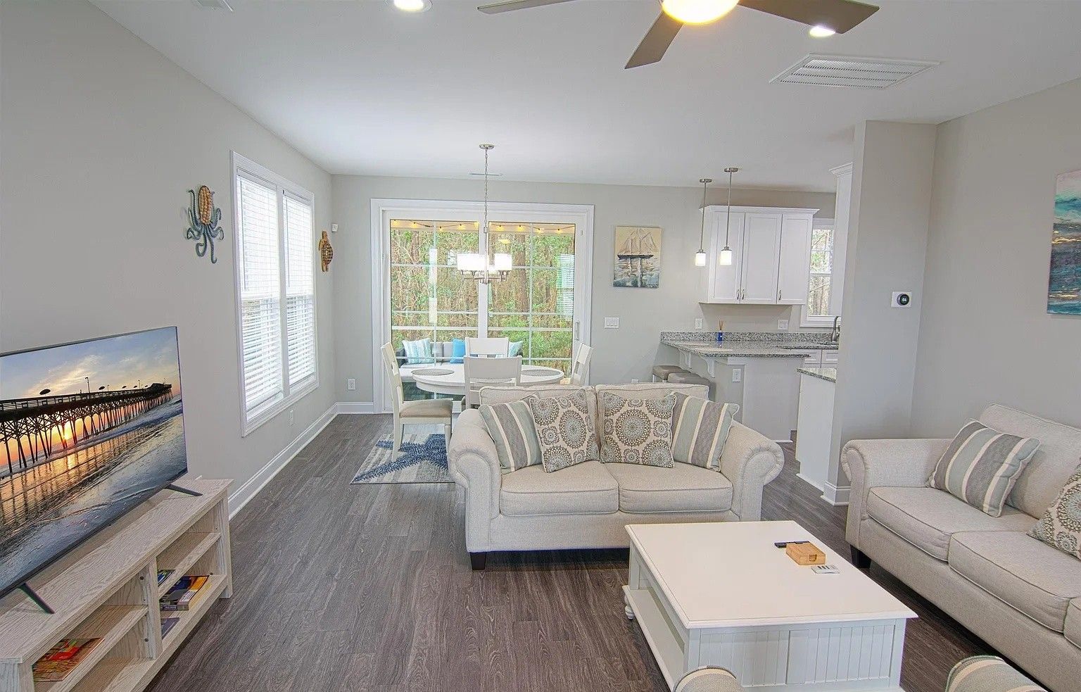 A modern living room with a sofa, coffee table, and TV. There is a dining area with a table and chairs. The room has large windows and a ceiling fan.