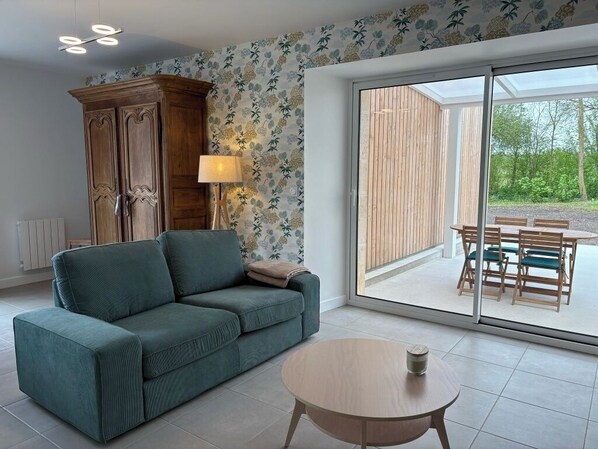 A living room with a green sofa, a wooden cabinet, a round coffee table, and a view of an outdoor patio with wooden furniture.