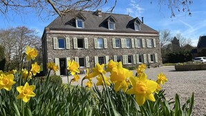 A stone building with multiple windows and shutters, surrounded by a gravel driveway and yellow daffodils.