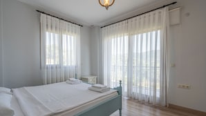Quarto com cama, mesa de cabeceira e janela com cortinas leves.