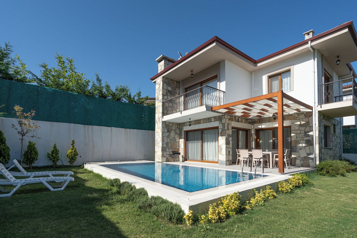 Une maison moderne à deux étages, dotée d’une piscine, d’un patio couvert et d’une façade en pierre.