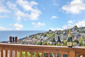 A balcony with a bottle of champagne and two glasses, overlooking a coastal town with houses and the sea.