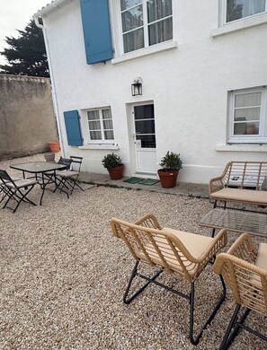 A white building with blue shutters, a gravel patio, wicker furniture, and potted plants.