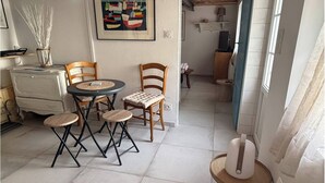 A dining area with a round table and four chairs, a white cabinet, a painting on the wall, and a window with curtains. - La voile blanche - A stopover with your feet in the water (Barbâtre)