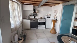 A compact kitchen with white cabinets, a microwave, stove, and a sink. There is a window with curtains, a door, and a small table with a basket. - La voile blanche - A stopover with your feet in the water (Barbâtre)