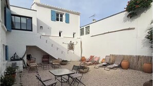 A courtyard with outdoor seating, a staircase, and a white building with blue shutters. - La voile blanche - A stopover with your feet in the water (Barbâtre)