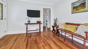A living room with hardwood flooring, a wooden bench with cushions, a small table with a basket and plant, and a flat-screen TV mounted on the wall. - Quaint Mill Cottage (Marion)