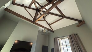 A room with exposed wooden beams, a hanging light fixture, a flat-screen TV mounted on the wall, and a window with curtains.