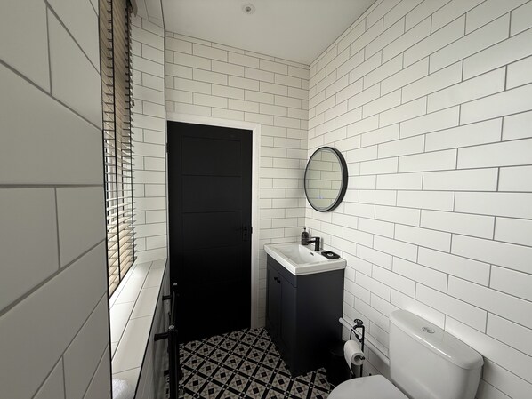 Un baño pequeño con paredes de azulejos blancos, una puerta negra, un lavabo blanco con un espejo encima y un inodoro.