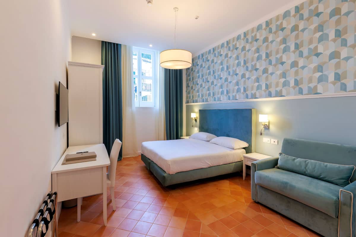 Habitación de hotel con cama, escritorio, sillón, sofá, televisor y ventana con cortinas, además de una pared con diseño.
