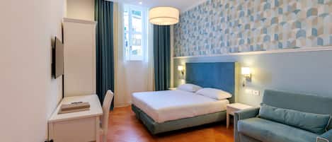 A hotel room with a bed, a desk, a chair, a sofa, a TV, a window with curtains, and a patterned wall.