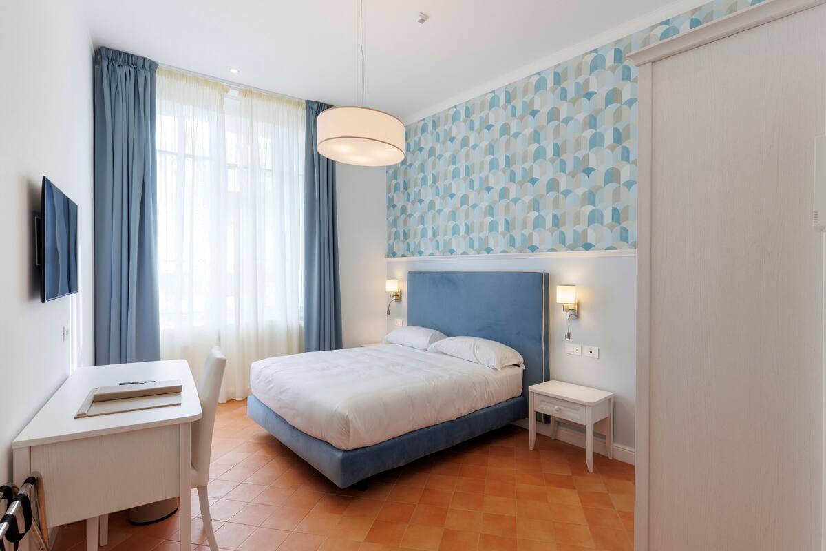 Una habitación de hotel moderna con cama, escritorio, silla, mesita de noche pequeña y televisor montado en la pared.